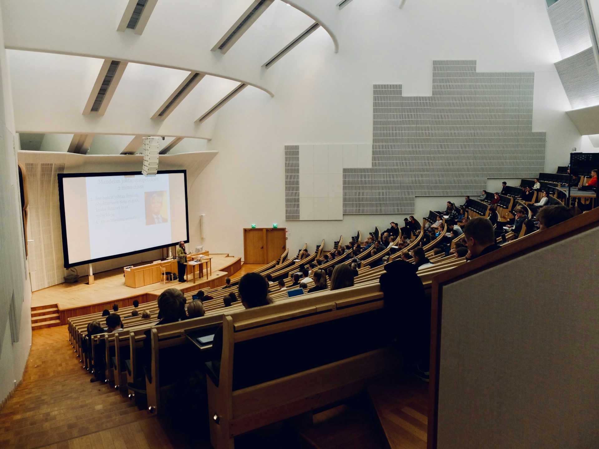 A packed university lecture hall with students seated in tiered wooden benches watching a professor present at the front, illustrating the freshman college classroom experience where GPA habits are formed.